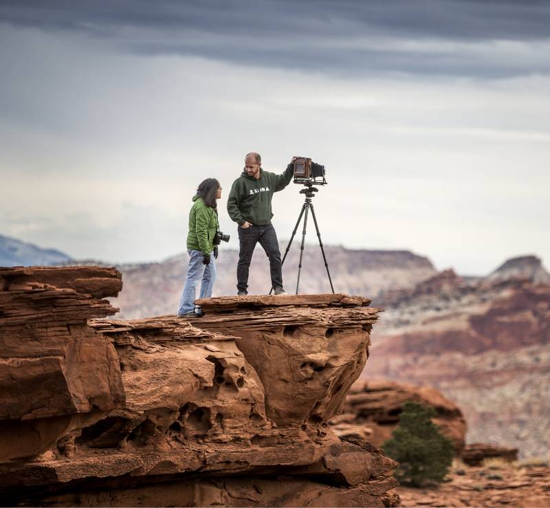 Two people on a red rock cliff, one adjusting a camera on a tripod