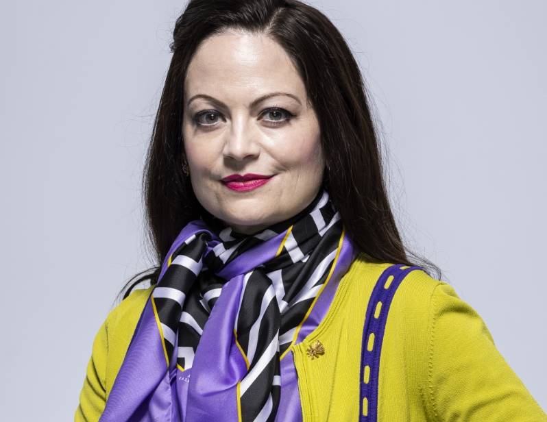 Woman wearing a yellow cardigan and mulicolored scarf against a gray background