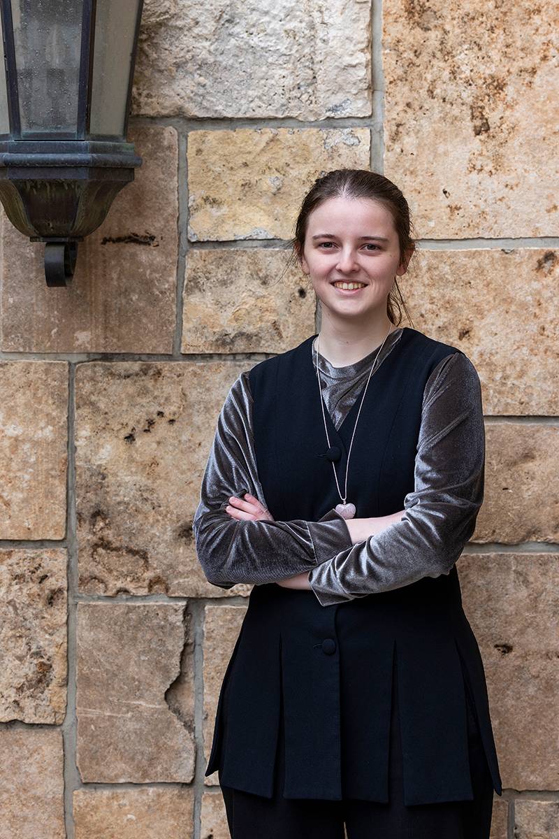 Person standing in front of a stone wall, wearing a velvet top and black vest, smiling with arms crossed.              