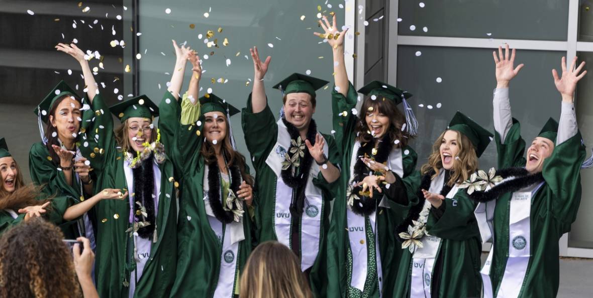 A group of graduates in green caps and gowns celebrating by tossing confetti in the air