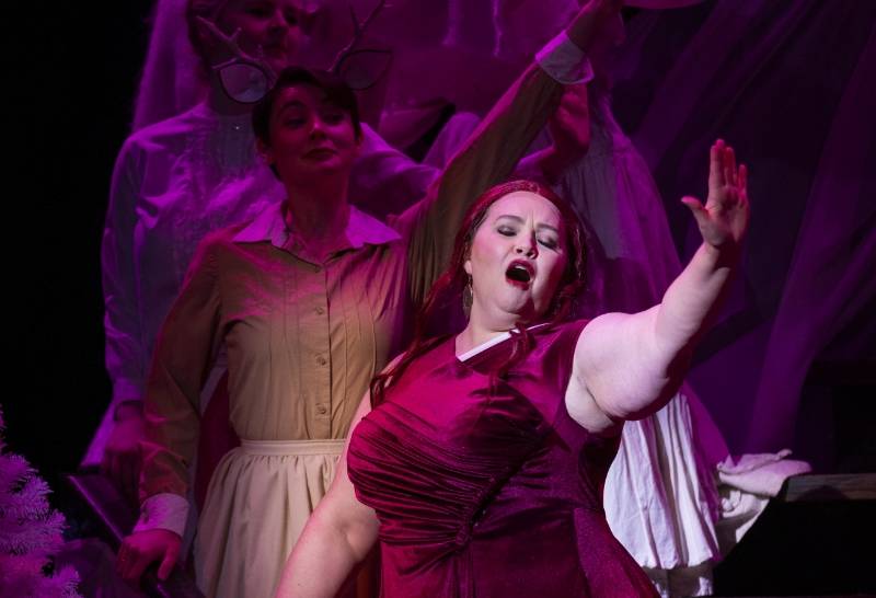 A women in a red dress sings on stage with other performers in the backgrounod under purple lighting